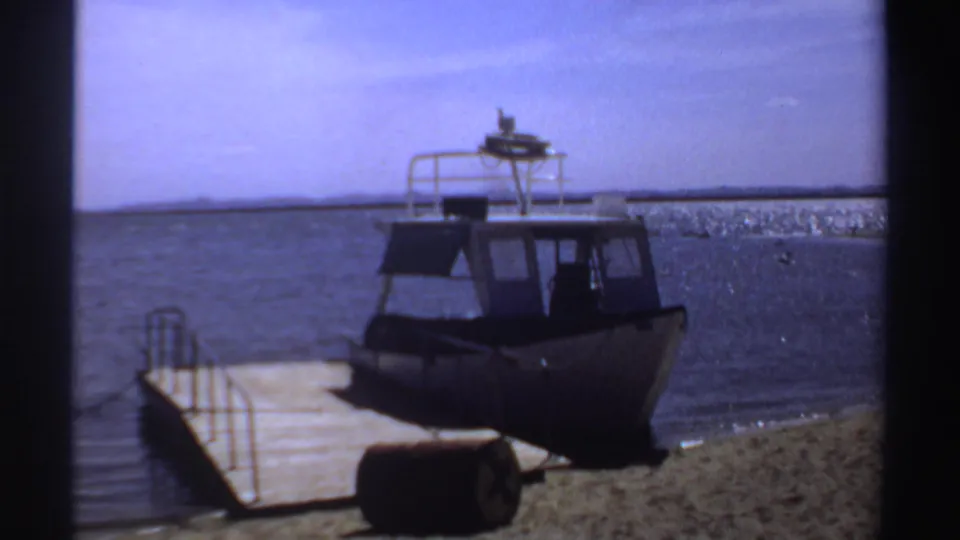 1969 Sudan waterfront frame showing a small motorboat tied near a wooden dock with open water beyond.
