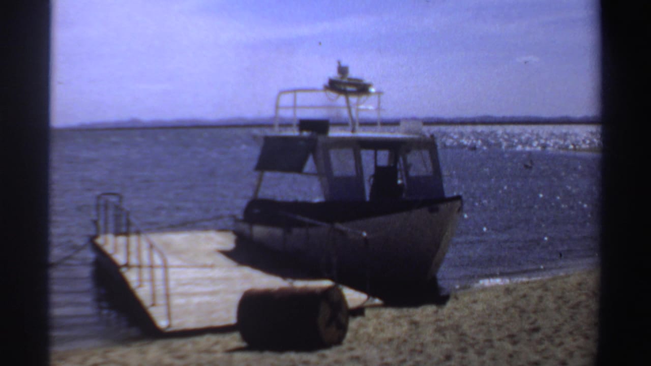 1969 Sudan 8mm frame showing a small cabin motorboat docked at a wooden pier beside a lagoon shoreline.