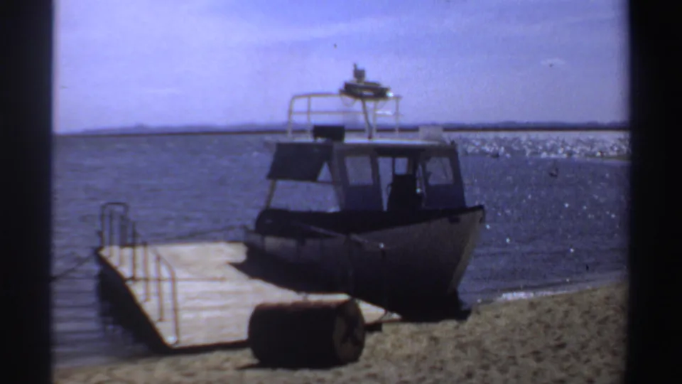 1969 Sudan 8mm frame showing a small cabin motorboat docked at a wooden pier beside a lagoon shoreline.