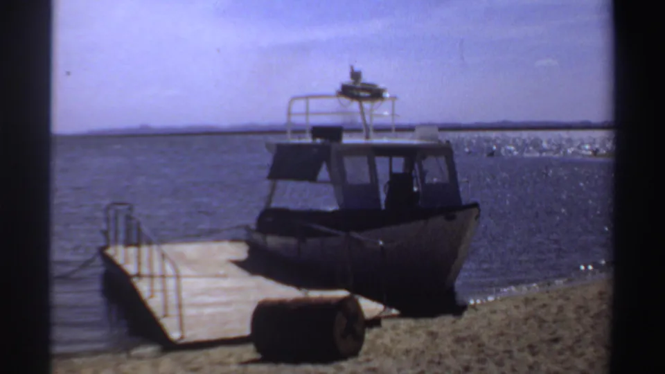 1969 Sudan 8mm frame showing a moored motorboat, wooden pier, sandy shore and dark barrel-like object in front.