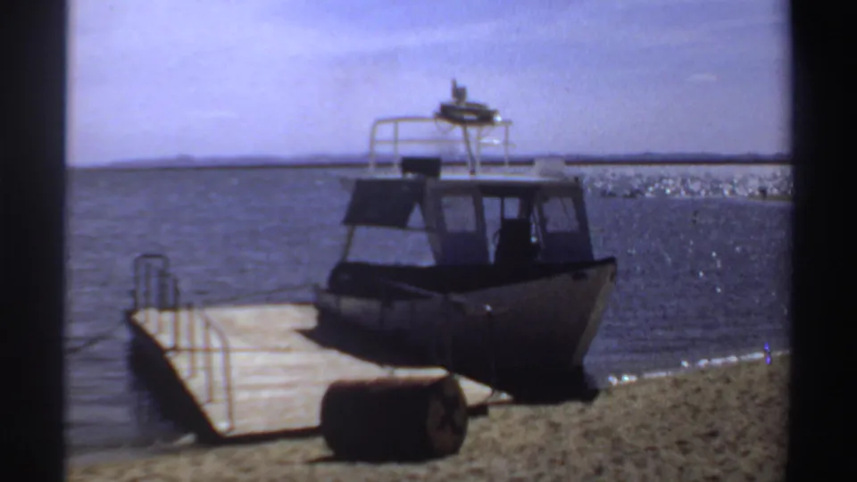 1969 Sudan 8mm frame of a stationary cabin motorboat at a wooden pier on a calm lagoon shore.