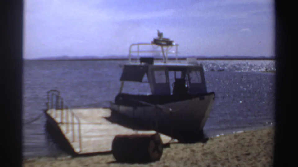 1969 Sudan home-movie frame showing a small powerboat beside a wooden dock on calm water near a sandy shoreline.