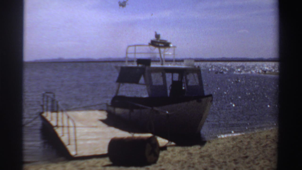 1969 Sudan frame of a moored small powerboat, its dark cabin and white roof rail set against sunlit water.