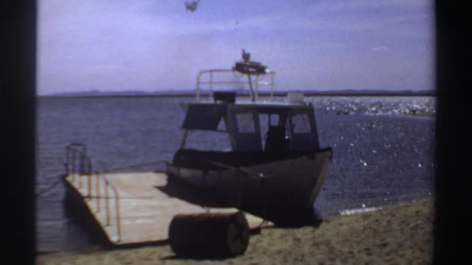 1969 Sudan frame of a moored small powerboat, its dark cabin and white roof rail set against sunlit water.