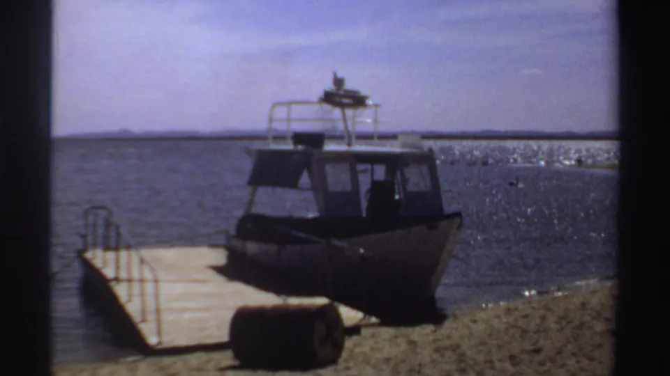 1969 Sudan home-movie view of the same powerboat from shore, with dock planks, sand, and calm open water visible.