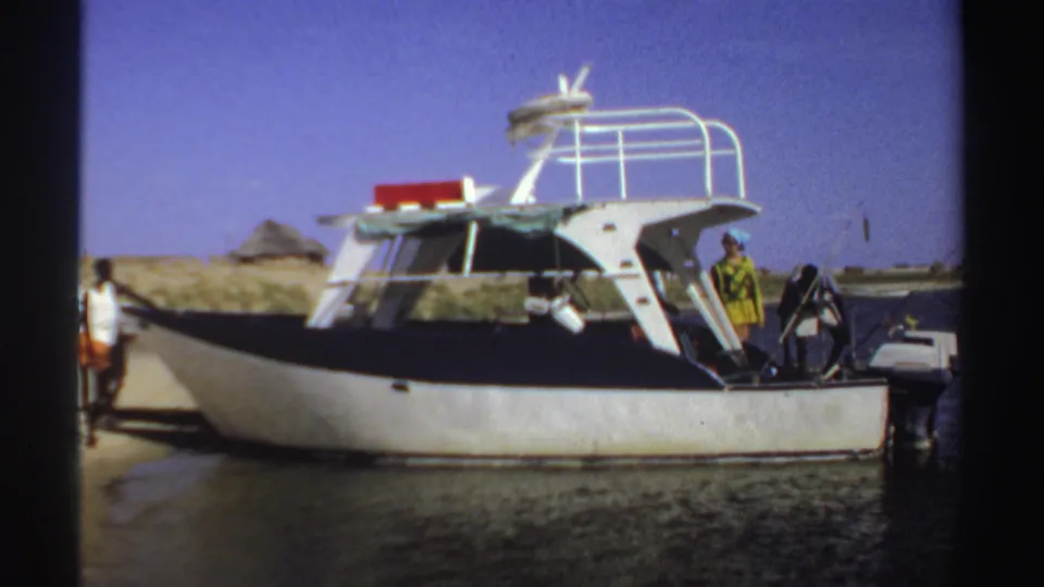 1969 Sudan frame showing a woman in a yellow dress on the deck of a small powerboat, with another person nearby on shore.