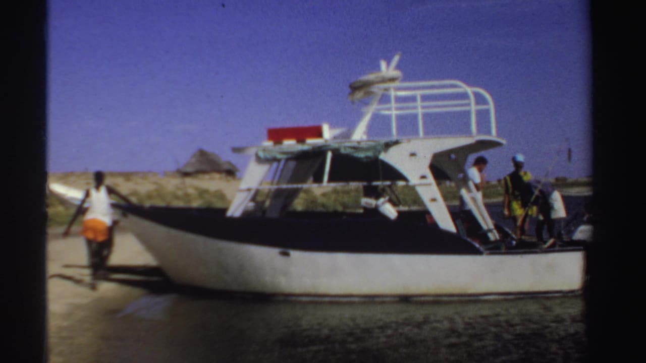 Motorboat beside shore as a person in orange shorts stands near the bow and others are at the stern.
