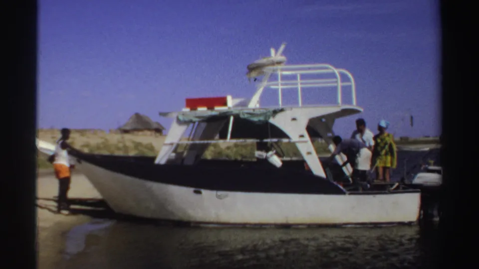 1969 Sudan home-movie frame showing people gathered around a white motorboat on a sandy shore under a clear blue sky.