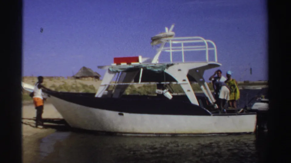 1969 Sudan frame showing people at the stern of a white motorboat, with sandy ground, water, and a thatched hut visible behind.