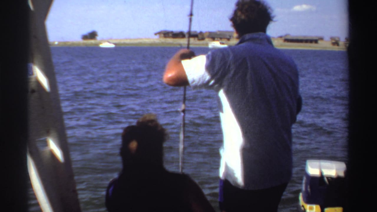 In Sudan home-movie footage from 1969, a man in a white shirt fishes from a boat with blue water and a low shoreline beyond.