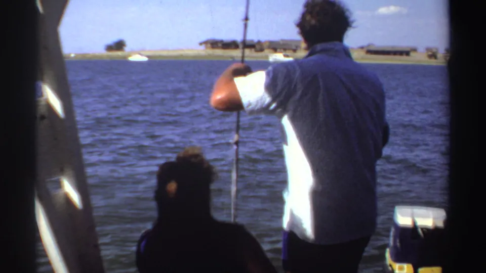 In Sudan home-movie footage from 1969, a man in a white shirt fishes from a boat with blue water and a low shoreline beyond.