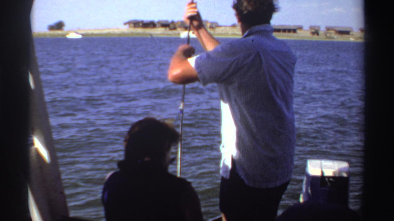 The fisherman stands with his back to the camera, rod raised over calm water, with Sudan shoreline structures visible in the distance.