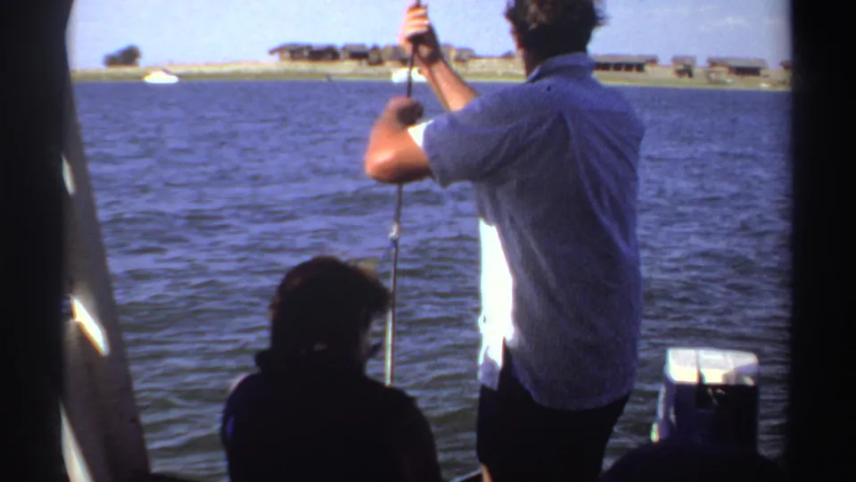 The fisherman stands with his back to the camera, rod raised over calm water, with Sudan shoreline structures visible in the distance.