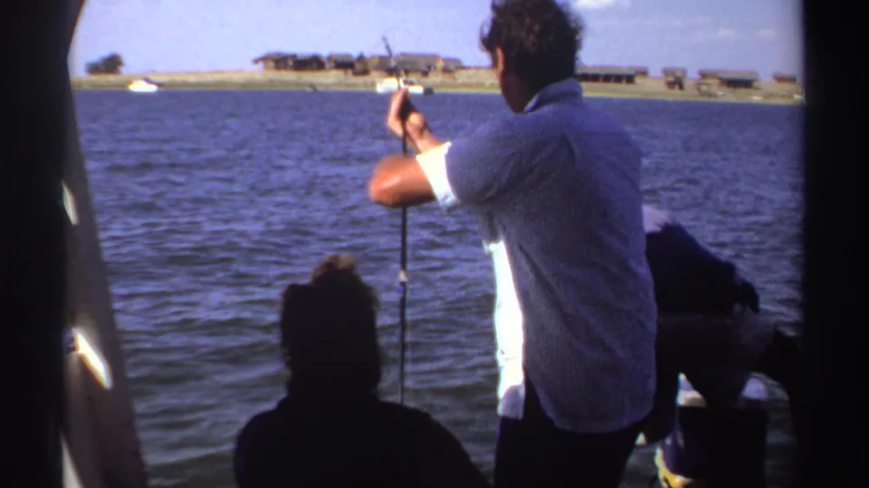 1969 Sudan home-movie frame of a man casting a fishing rod from a boat, with seated passengers and a distant shoreline across the water.