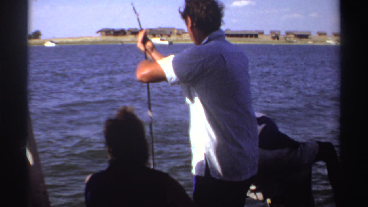 1969 Sudan frame showing a man fishing from a boat, with dark passenger silhouettes, blue water, and low shoreline structures in the distance.