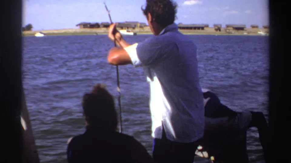 1969 Sudan frame showing a man fishing from a boat, with dark passenger silhouettes, blue water, and low shoreline structures in the distance.