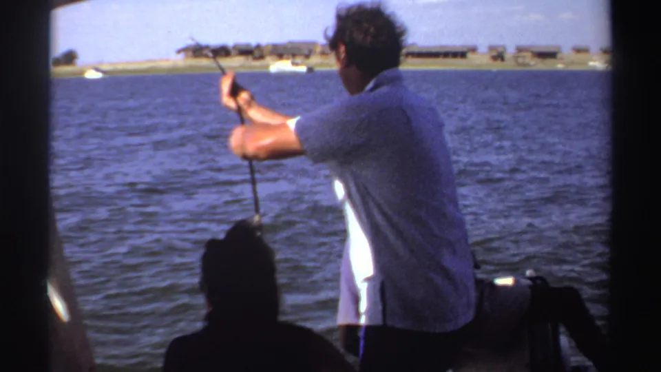1969 Sudan home-movie still of a man fishing from a boat, framed by dark boat edges and seated passengers against a wide body of water.
