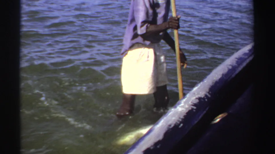 1969 Sudan frame showing a person standing in shallow water near the boat with a long pole, with rippling water and boat rail visible.