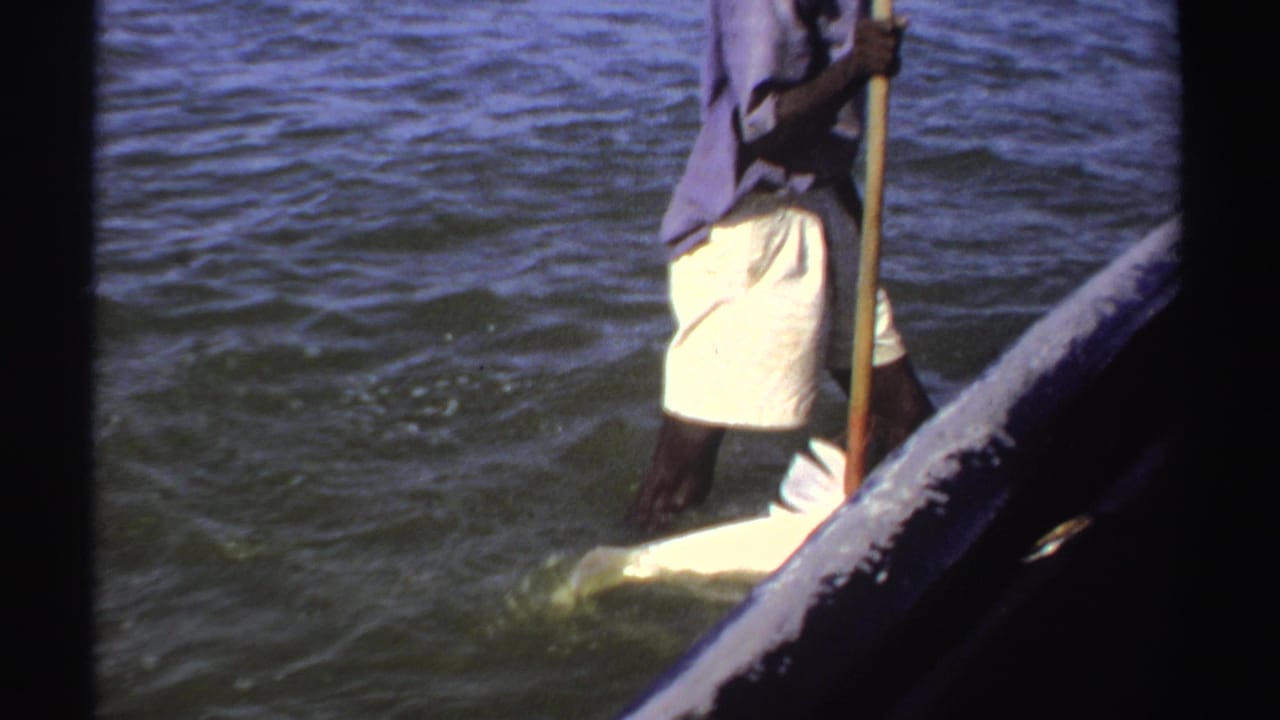 1969 Sudan 8mm frame showing a man in shallow water beside a small boat, using a long pole near a large pale fish.
