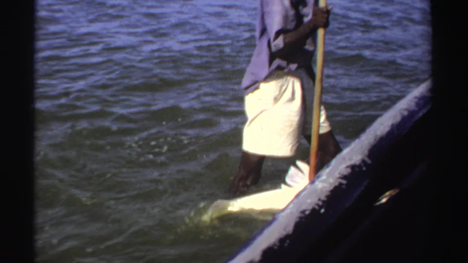 1969 Sudan 8mm frame showing a man in shallow water beside a small boat, using a long pole near a large pale fish.