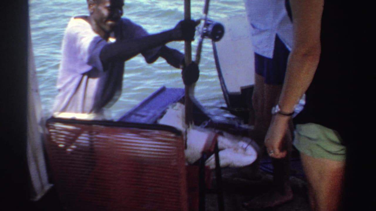 1969 Sudan 8mm frame showing adults on a small boat, water in the background, and a man in a white shirt near boat gear and a catch.