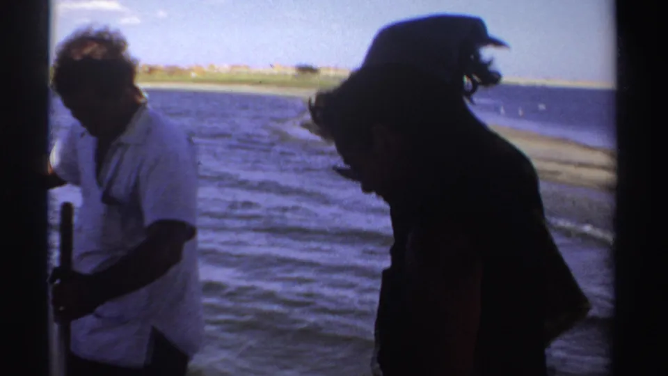 1969 Sudan 8mm frame showing a man in a white shirt and another adult silhouetted on a boat, with open water and shoreline in the background.