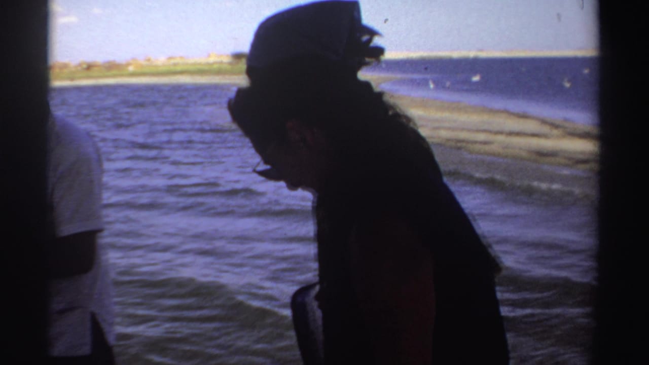 1969 Sudan home-movie frame of adults on a boat, one in a dark hat silhouetted against bright water and shoreline.