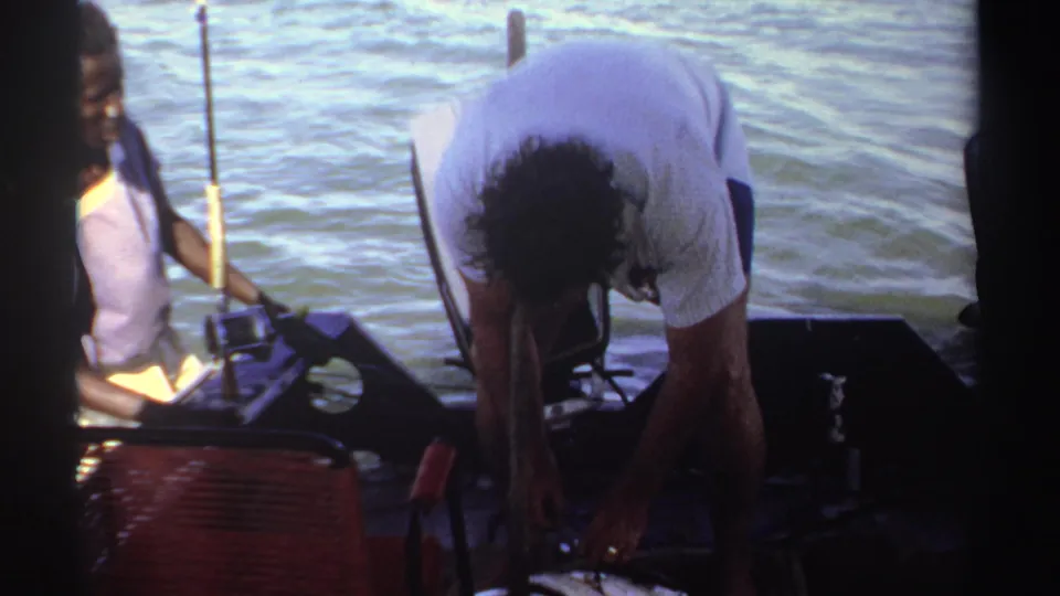 1969 Sudan home-movie frame of a person bending over gear on a boat deck, with another adult seated by the water.