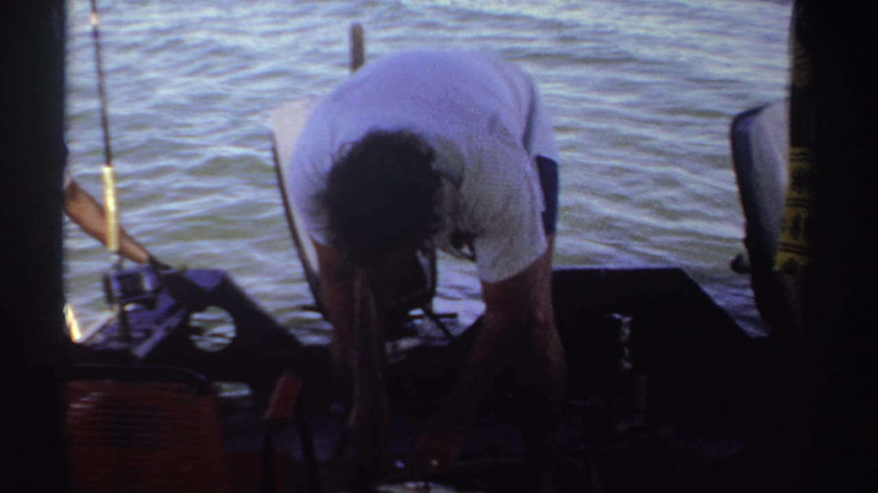 Man bends over a large fish on a boat deck beside fishing gear and open water.