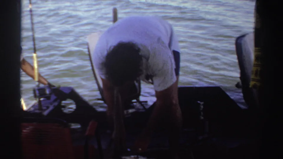 In 1969 Sudan home-movie footage, a man in a white shirt bends over a large fish on a boat deck near rods and calm water.