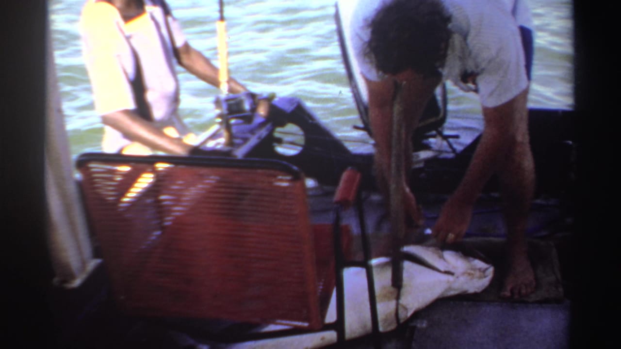 1969 Sudan boat-deck scene with one man holding a fishing rod as another bends near a large fish beside a red woven chair.