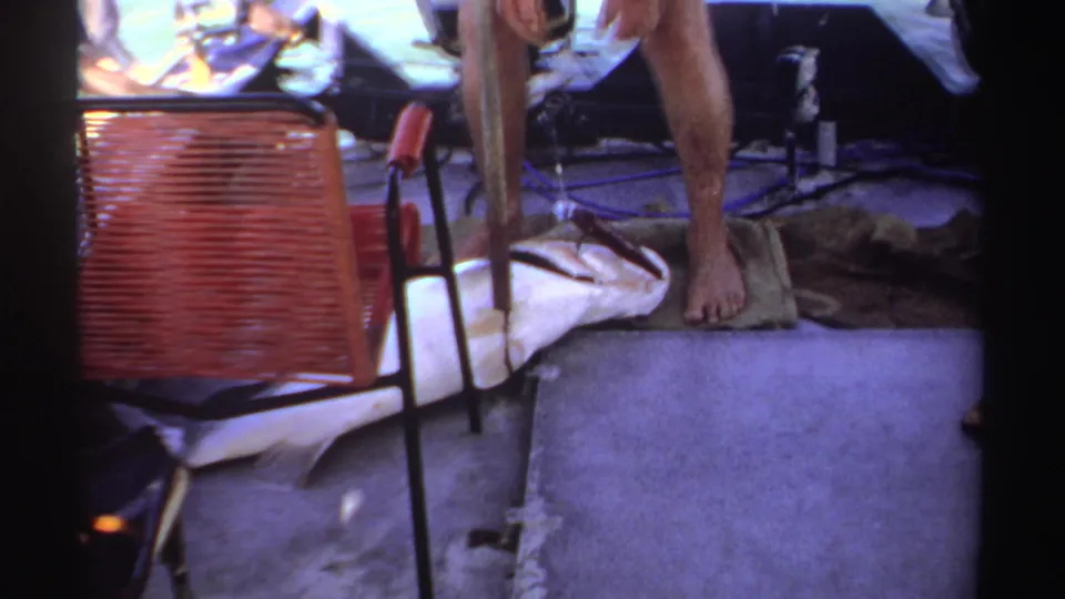 A barefoot fisherman in 1969 Sudan footage stands over a large fish on the boat deck near towels, tackle, and a red woven chair.