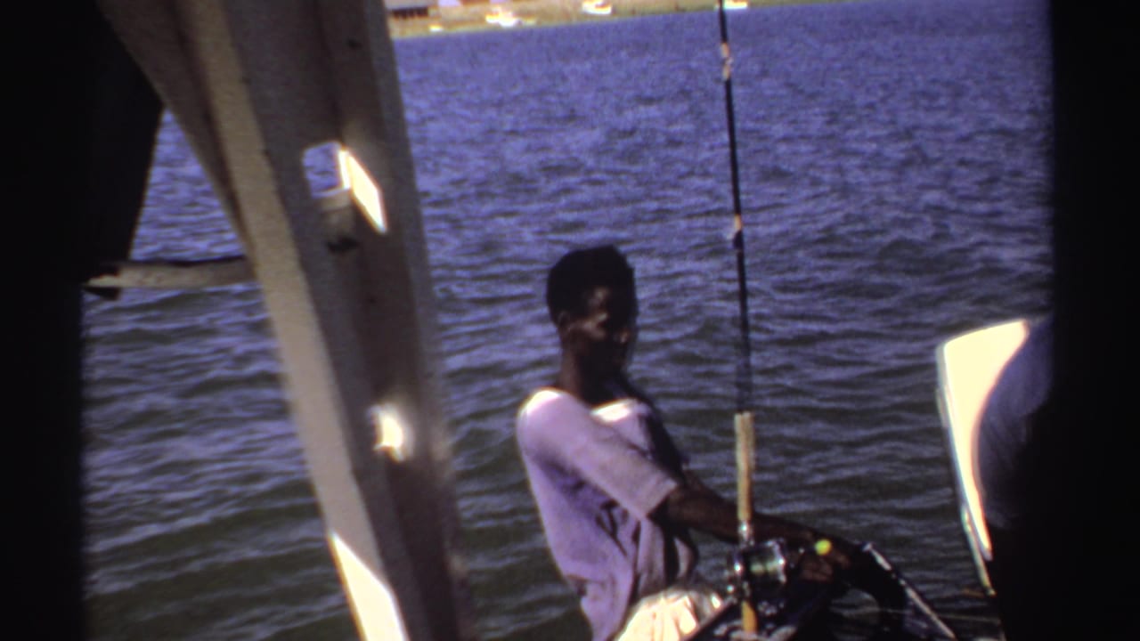A man sits or leans near a fishing rod on a small boat in Sudan-associated 1969 home-movie footage, with calm water behind him.