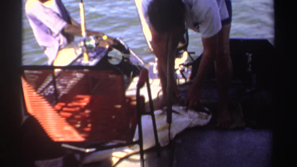 A man bends down over a large fish on the deck of a small boat, with a red woven chair and fishing gear nearby.
