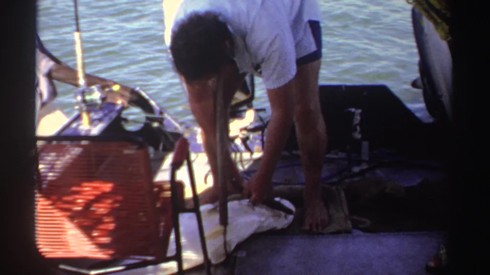 A barefoot man in a light shirt bends over the fish on the boat deck, with rods, a red chair, and water visible around him.