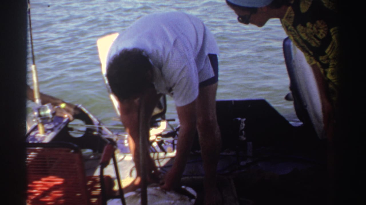 Man bends over fishing gear on a small boat while a woman stands nearby on open water.