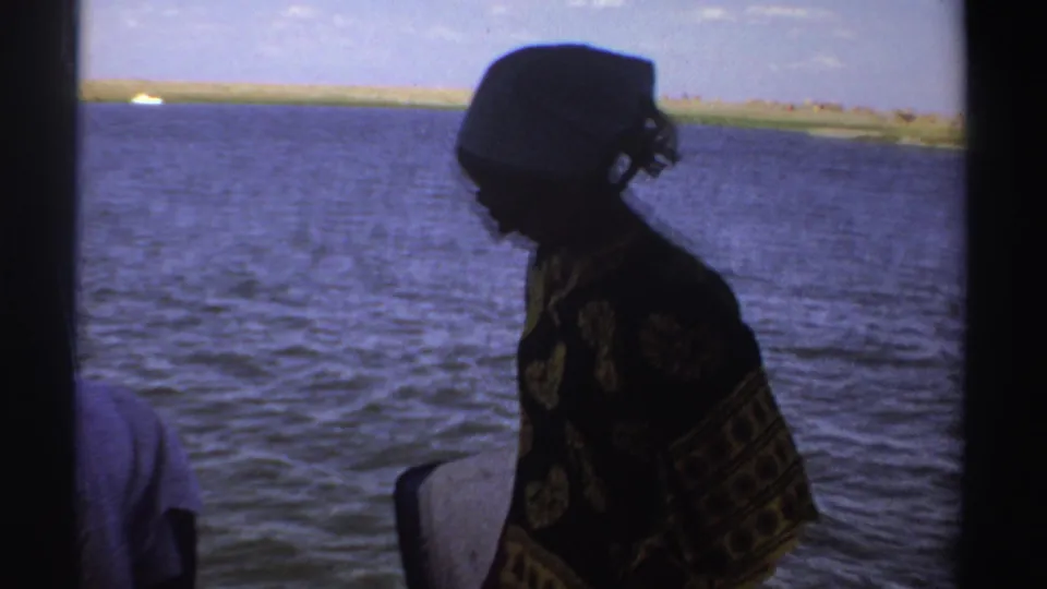 A woman in a patterned dress and white headscarf stands in profile on a boat, with open water and a low shoreline behind her in Sudan.
