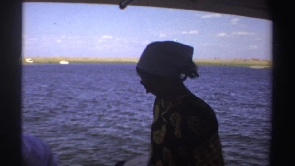 The woman remains on the boat in profile, her white headscarf silhouetted against water and a low Sudan shoreline.