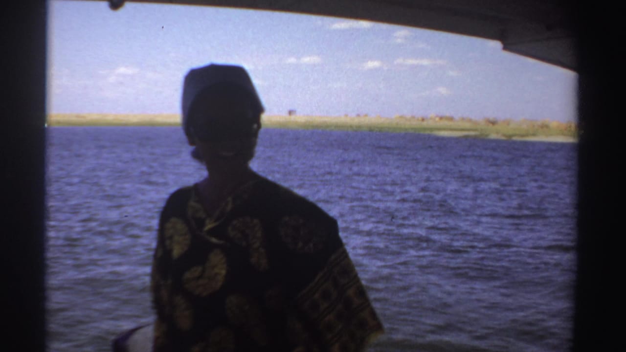Silhouetted person in patterned shirt and hat stands on a small boat beside blue water.