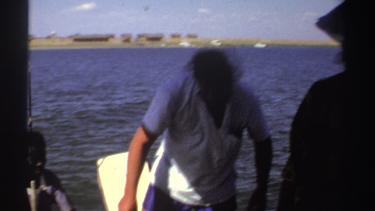 A person in a light shirt leans near the motor area of a small boat on Sudanese river water, with another figure partly visible.