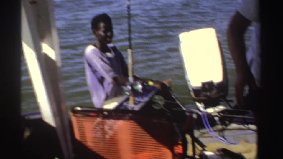 A seated passenger on a small Sudan motorboat sits near fishing rods and equipment, framed by water and bright sunlight.