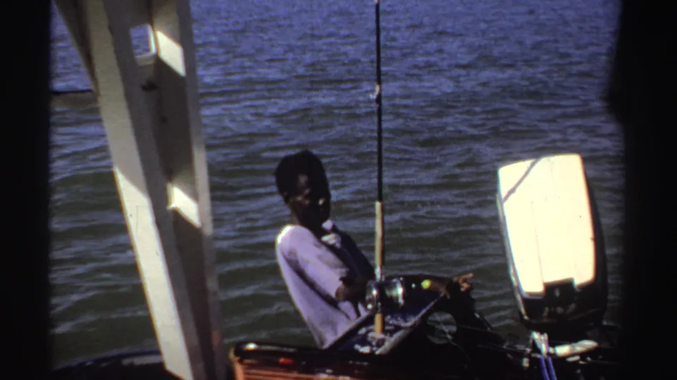 A seated passenger remains near fishing rods on a small motorboat in Sudan, with rippled river water filling most of the frame.
