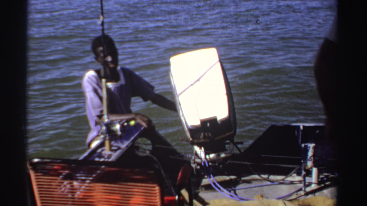 Adult man seated in a small boat beside water and a bright rectangular panel.