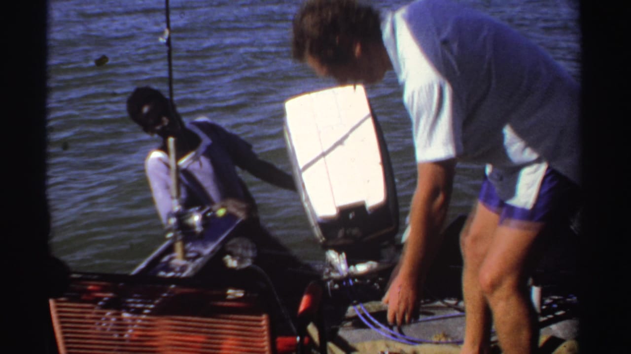 In 1969 Sudan home-movie footage, a seated man and a bending assistant are visible on a small boat beside open water.