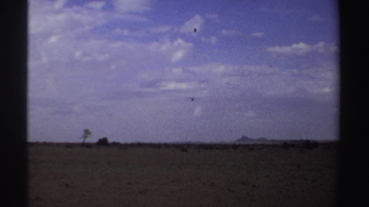 Small airplane above a wide brown field under a blue sky with scattered clouds.