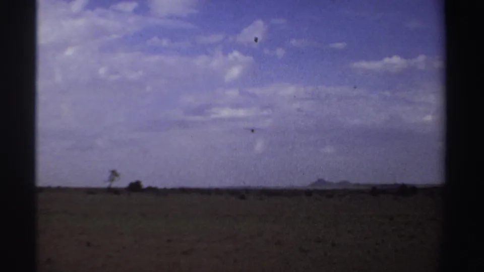1969 Sudan 8mm frame showing a small airplane over a dry open field, with sparse vegetation and distant low hills.
