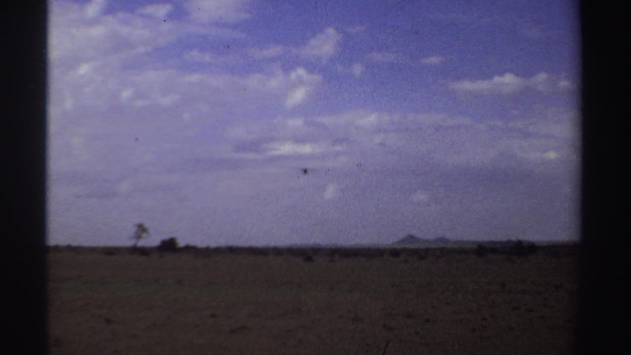 Small airplane flying across a distant horizon over a dry field and cloudy sky.