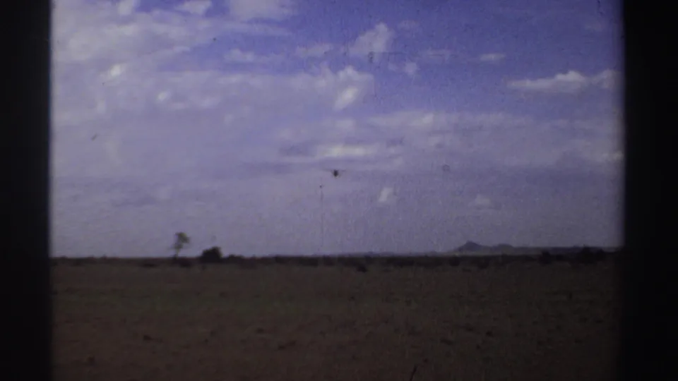 1969 home-movie frame of a small airplane over an open Sudan field, with distant hills and visible film grain.