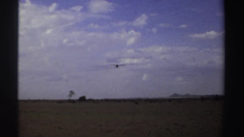 A low aircraft crosses a dry Sudanese field in a faded 8mm frame, with sparse vegetation and hills on the horizon.
