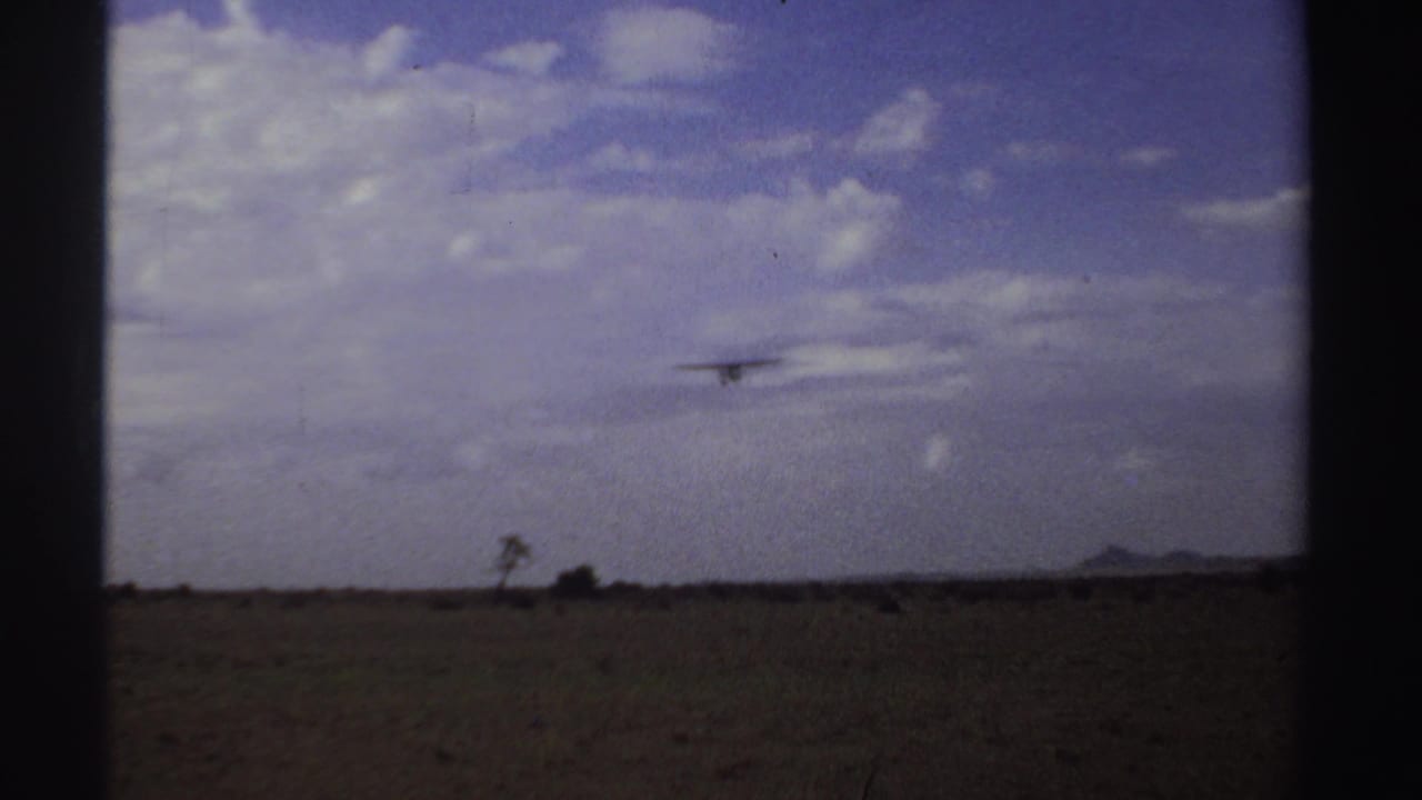 Small airplane distant over flat dry terrain beneath blue sky and grey clouds.